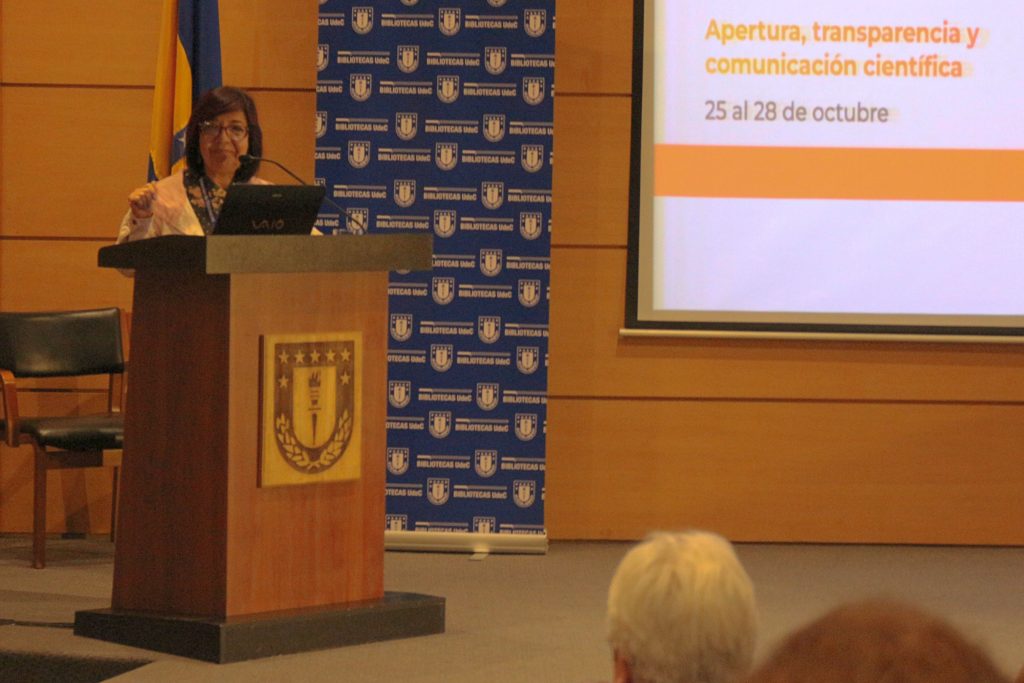 Doctora Lourdes Feria expone desde un podio con el logo de la Universidad de Concepción, al fondo hay una pantalla donde se proyecto donde se lee: apertura, transparencia y comunicación científica.