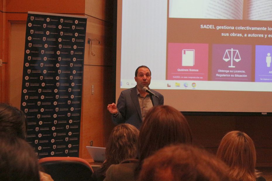 El Director de la Sociedad del Derecho de Autor, Cristian Elgueta, habla en un micrófono ubicado delante de una pantalla donde se proyecta un Power Point.
